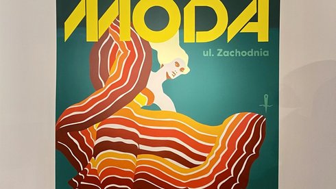 - W sali wystawowej wisi wielkoformatowa reprodukcja, a na niej wizerunek kobiet w kolorej sukni. Na górze obrazu napis "MODA". W sali wystawowej wisi wielkoformatowa reprodukcja, a na niej wizerunek kobiet w kolorej sukni. Na górze obrazu napis "MODA".